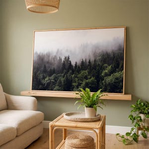 May include: A framed print showcases a misty forest scene with evergreen trees. The artwork is mounted above a wooden shelf, alongside a rattan side table with a plant and a woven lampshade.