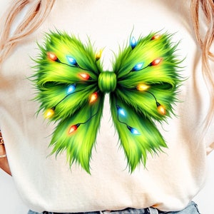 May include: Cream-colored t-shirt featuring a large, vibrant green bow design adorned with colorful Christmas lights. The bow has a fluffy, textured appearance. The shirt is paired with light-wash blue jeans.