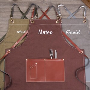May include: Three personalized aprons in olive green, brown, and gray. Each apron features a name embroidered in white: 'And', 'Mateo', and 'David'. The brown apron has a leather pocket with a fork and knife. The aprons have adjustable straps and metal hardware.