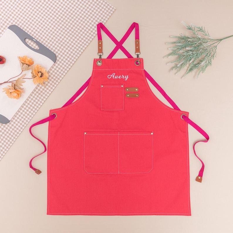 May include: A bright red apron with the name "Avery" embroidered on the chest. The apron has two large pockets and a smaller pocket. The straps are pink and brown. The apron is made of a sturdy fabric, perfect for cooking or crafting.