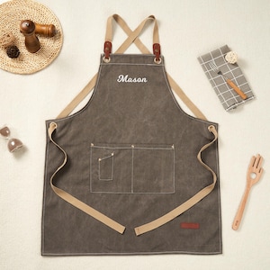 May include: A grey apron with the name "Mason" embroidered in white. It features tan straps and pockets, with leather details. Suitable for kitchen or workshop use.