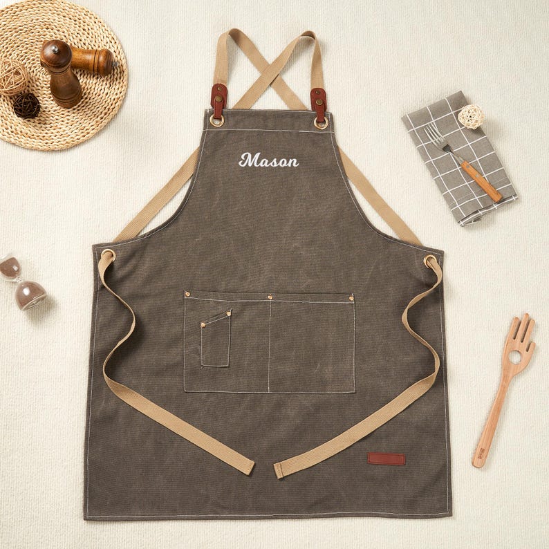 May include: A grey apron with the name "Mason" embroidered in white. It features tan straps and pockets, with leather details. Suitable for kitchen or workshop use.