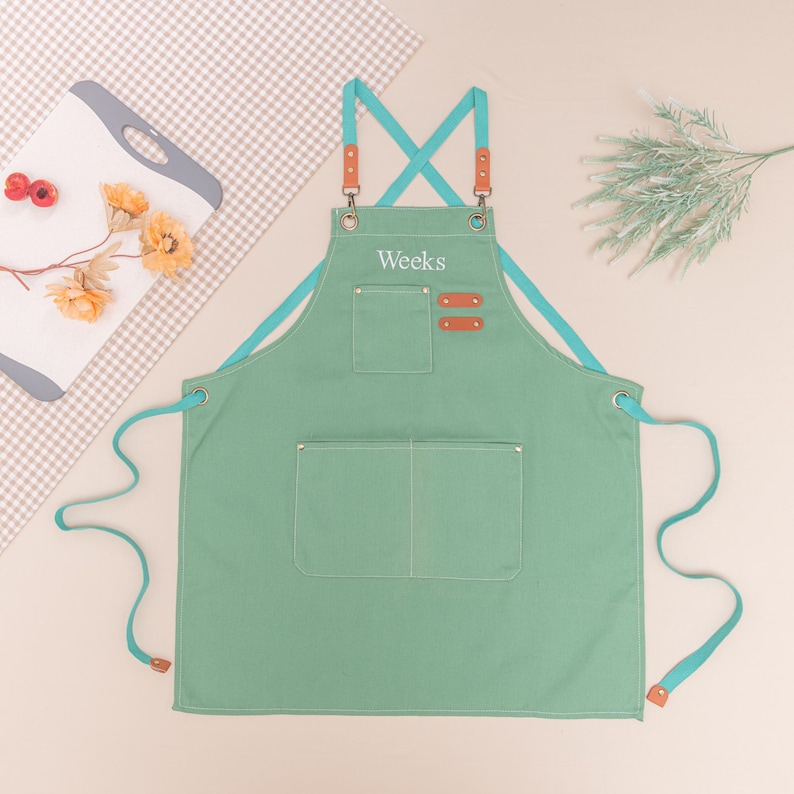 May include: A light green apron with teal straps and accents. The apron has two front pockets and leather-like patches. The word "Weeks" is embroidered on the chest. The apron is designed for kitchen or craft use.