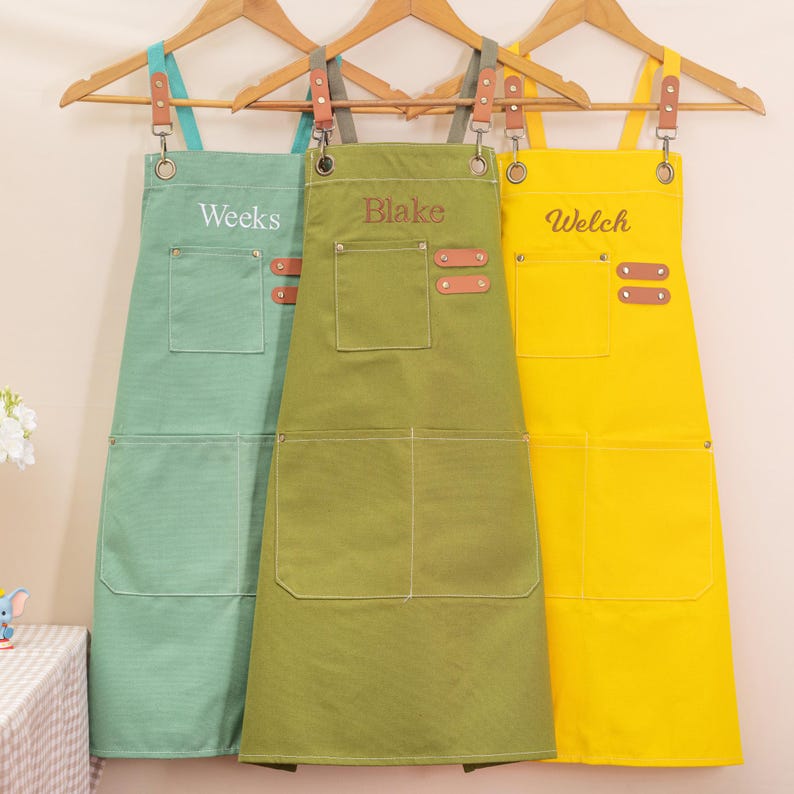 May include: Three personalized aprons in shades of green and yellow hang on wooden hangers. Each apron has a large front pocket and leather accents. The names "Weeks", "Blake", and "Welch" are embroidered on the aprons.