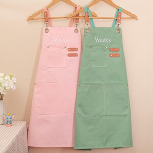 May include: Two aprons hanging on wooden hangers. One is light pink with the name "Lynne" embroidered on it, and the other is light green with the name "Weeks" embroidered on it. Both aprons have pockets and leather accents.