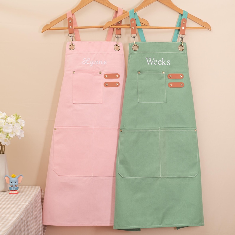 May include: Two aprons hanging on wooden hangers. One is light pink with the name "Lynne" embroidered on it, and the other is light green with the name "Weeks" embroidered on it. Both aprons have pockets and leather accents.