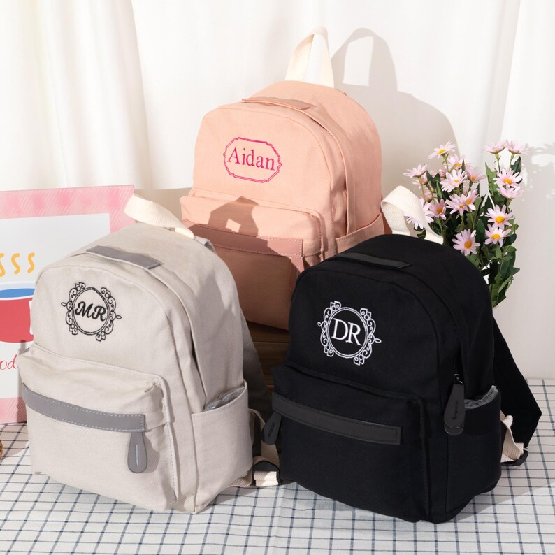 Puede incluir: Tres mochilas en beige, rosa claro y negro. La mochila beige presenta un monograma circular con las letras MR. La mochila rosa muestra el nombre Aidan en un marco decorativo. La mochila negra tiene un monograma circular con las letras DR.