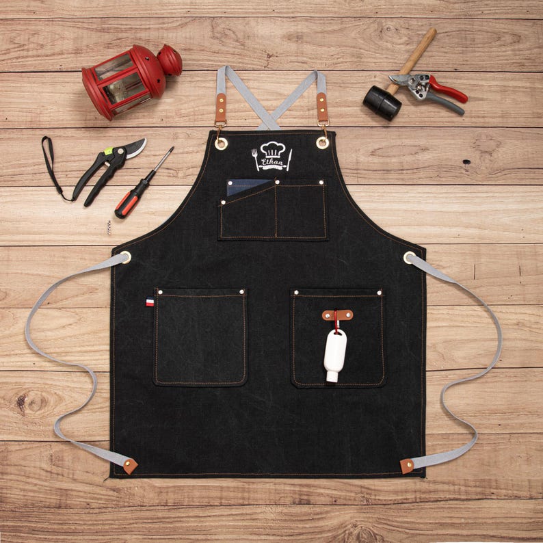 Peut inclure: Tablier noir avec plusieurs poches et des sangles grises, avec un logo de toque de chef et le nom "Tihaa". Le tablier a des accents en cuir marron et est entour&eacute; d'outils de jardinage et d'une lanterne rouge.