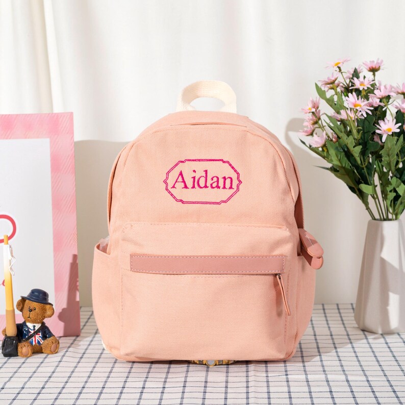 Puede incluir: Una mochila de lona color melocot&oacute;n claro con un asa superior y un bolsillo frontal con cremallera. El centro de la mochila muestra el nombre Aidan bordado en color rosa oscuro dentro de un marco rectangular decorativo con bordes festoneados.