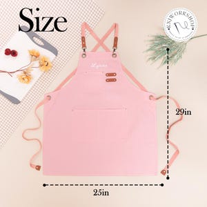 May include: A pink apron with the name "Lynne" embroidered on the chest. The apron has two pockets and brown leather accents. It measures 29 inches in length and 25 inches in width.