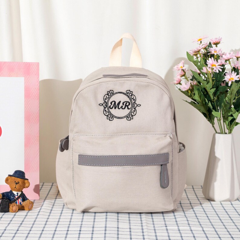 Puede incluir: Una mochila de lona color beige claro con un asa superior y un bolsillo frontal con cremallera y ribete gris. El centro de la mochila muestra un emblema circular bordado en negro con adornos decorativos y las letras MR en el centro.