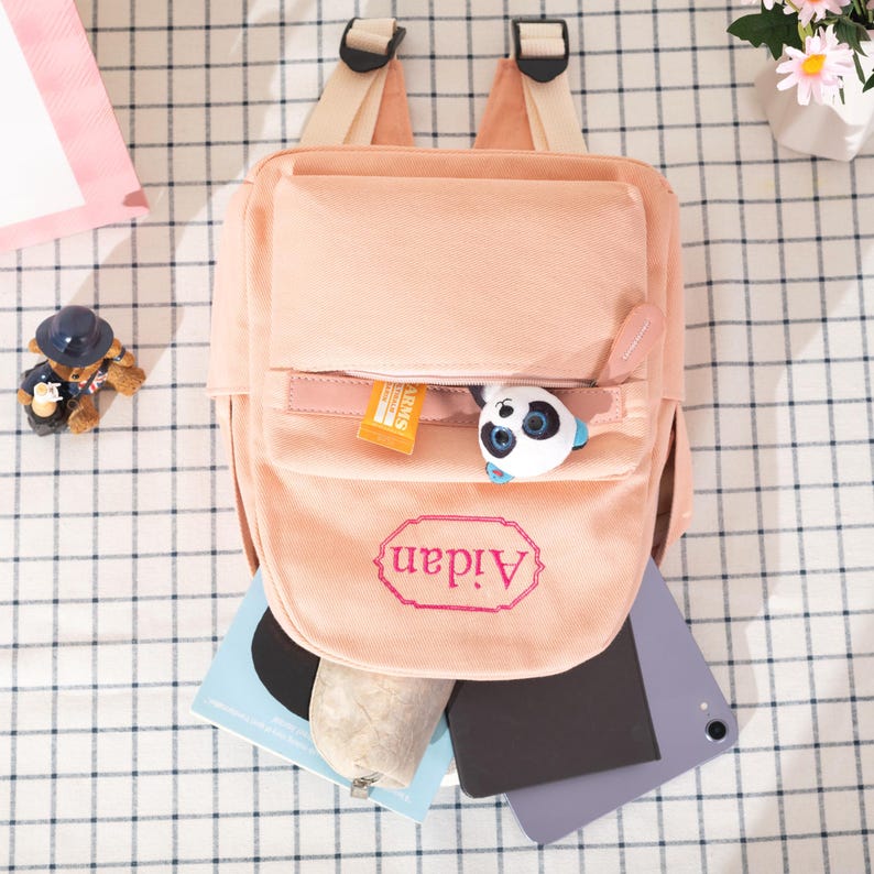 Puede incluir: Una mochila de lona color melocot&oacute;n con el nombre Aidan bordado en rosa dentro de un borde decorativo. El bolsillo delantero est&aacute; parcialmente abierto, revelando un peque&ntilde;o peluche de panda y un paquete naranja dentro del compartimento con cremallera.
