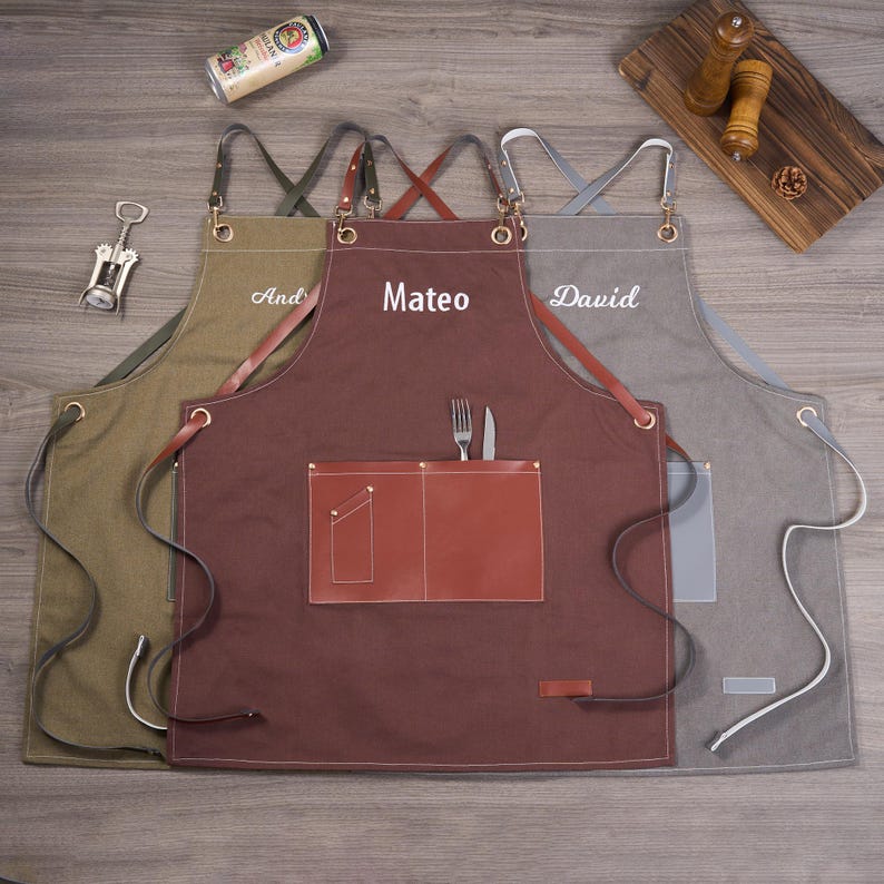 May include: Three personalized aprons in olive green, brown, and gray. Each apron features a name embroidered in white: 'And', 'Mateo', and 'David'. The brown apron has a leather pocket with a fork and knife. The aprons have adjustable straps and metal hardware.