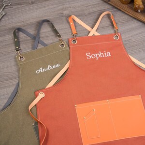 May include: Two personalized aprons. One is olive green with the name "Andrew" embroidered in white. The other is orange with the name "Sophia" embroidered in white. Both aprons have leather straps and pockets.