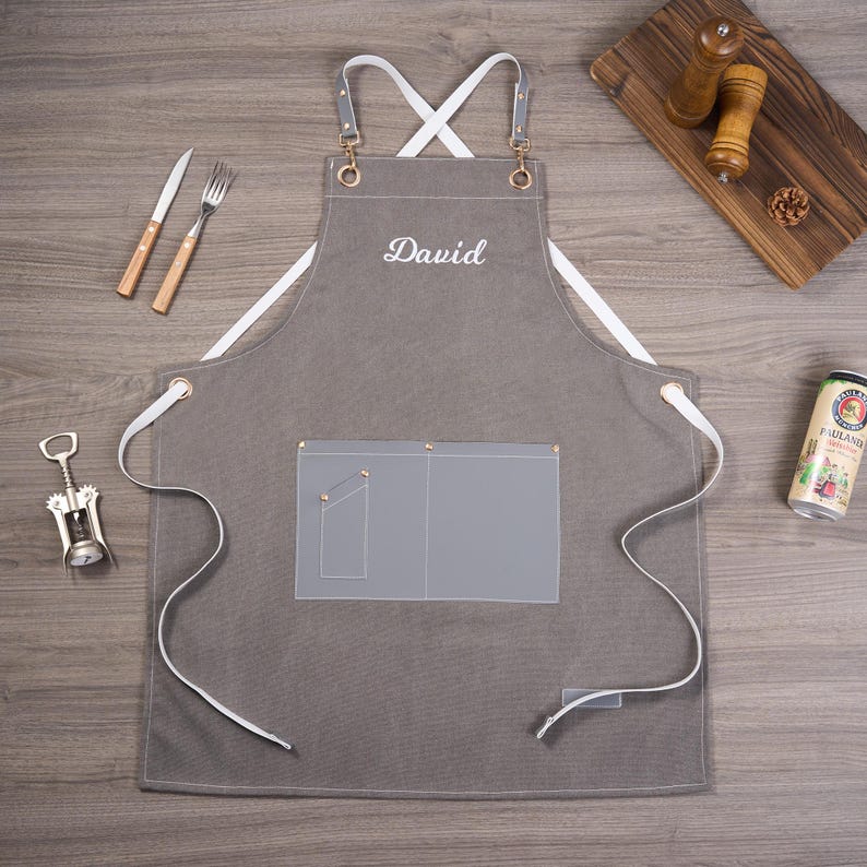 May include: Gray apron with white straps and the name "David" embroidered in white. Features a front pocket and metal grommets. Includes a fork, knife, and corkscrew. A beer can and wooden cutting board are also visible.