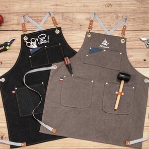 Peut inclure: Deux tabliers, un noir et un gris, avec plusieurs poches et des sangles grises. Le tablier noir porte le nom "Ethan" et un logo de toque de chef brod&eacute;. Le tablier gris porte le nom "Rowan" et un logo de flamme brod&eacute;.