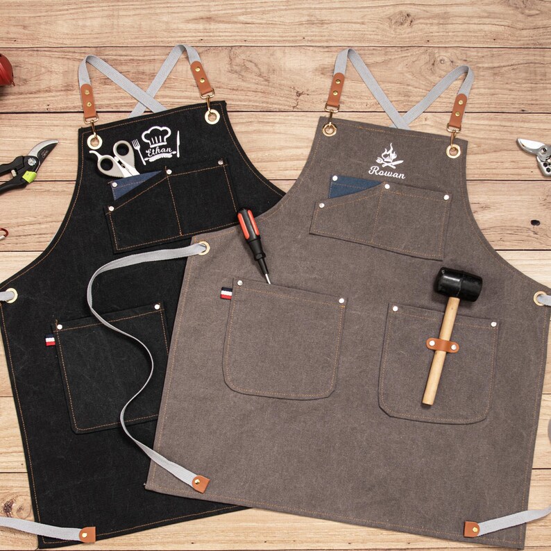 Peut inclure: Deux tabliers, un noir et un gris, avec plusieurs poches et des sangles grises. Le tablier noir porte le nom "Ethan" et un logo de toque de chef brod&eacute;. Le tablier gris porte le nom "Rowan" et un logo de flamme brod&eacute;.