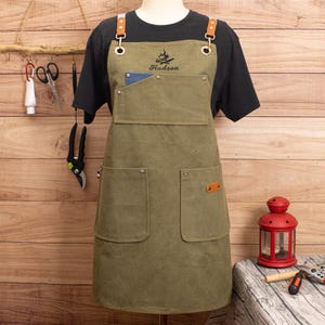 Peut inclure: Tablier vert olive avec des bretelles et des poches en cuir. Le tablier porte le nom "Hudson" brod&eacute; au-dessus d'un motif de feu de camp. Le tablier est pr&eacute;sent&eacute; sur un mannequin, avec un t-shirt noir en dessous.