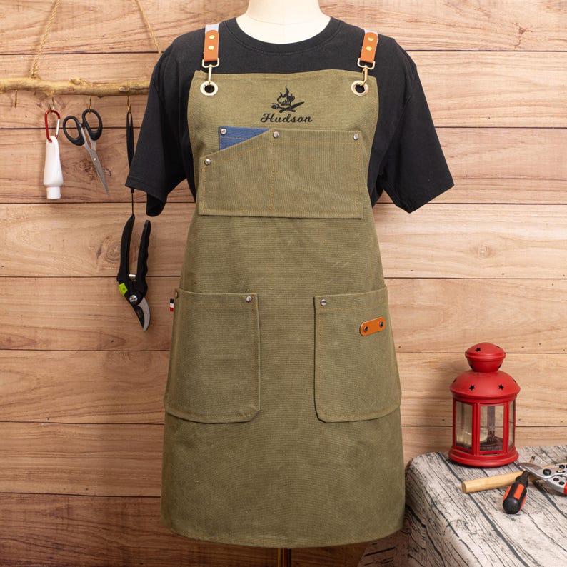 Peut inclure: Tablier vert olive avec des bretelles et des poches en cuir. Le tablier porte le nom "Hudson" brod&eacute; au-dessus d'un motif de feu de camp. Le tablier est pr&eacute;sent&eacute; sur un mannequin, avec un t-shirt noir en dessous.