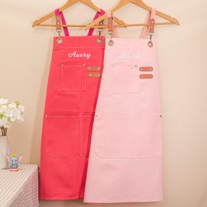 May include: Two aprons hanging on wooden hangers. One is a vibrant red with the name "Avery" embroidered in white. The other is a soft pink with "Lynne" embroidered in white. Both aprons have pockets and leather accents.