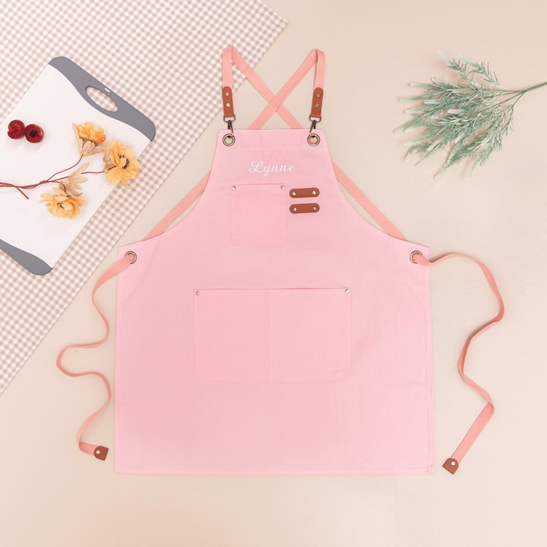 May include: A light pink apron with the name "Lynne" embroidered on the front. The apron has two pockets and brown leather accents on the straps. The apron is made of a soft fabric and has a cross-back design.