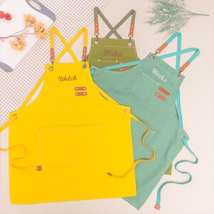 May include: Three aprons in yellow, green, and olive green with leather accents. The yellow apron has "Welch" embroidered on it. The green apron has "Weeks" embroidered on it, and the olive green apron has "Blake" embroidered on it. All aprons have pockets.