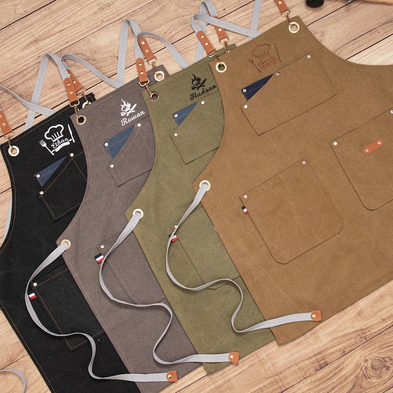 Peut inclure: Rang&eacute;e de tabliers personnalis&eacute;s noirs, gris, vert olive et marron. Chaque tablier porte un nom ou un logo unique, plusieurs poches et des d&eacute;tails en cuir et en m&eacute;tal. Les tabliers ont des sangles de cou et des liens de taille r&eacute;glables.