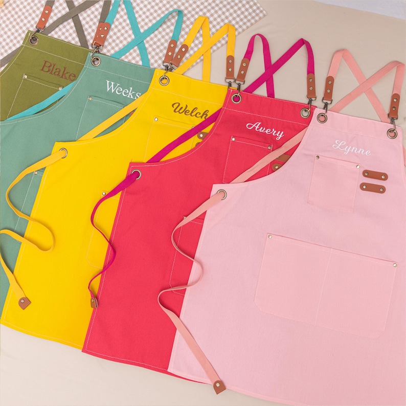 May include: A collection of colorful aprons in shades of green, yellow, red, and pink. Each apron features a pocket and personalized name embroidery. The aprons have adjustable straps with leather accents and metal hardware.