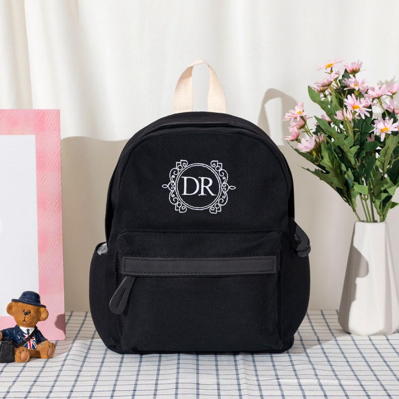 Puede incluir: Una mochila negra que presenta un emblema circular bordado en blanco con las letras DR en el centro. La bolsa tiene un asa superior, un bolsillo frontal con cremallera y una correa horizontal, adem&aacute;s de bolsillos laterales en el dise&ntilde;o.