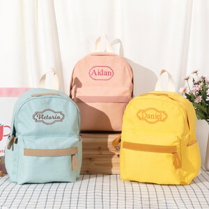 Puede incluir: Tres mochilas en azul claro, melocot&oacute;n y amarillo, cada una con un asa superior, un bolsillo frontal con cremallera y una correa beige, y un marco decorativo bordado con los nombres Victoria, Aidan y Daniel respectivamente.