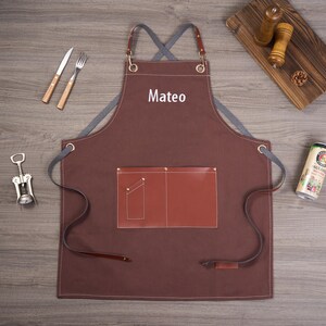 May include: A brown apron with the name "Mateo" in white, featuring a brown leather pocket and straps. The apron is on a wooden surface, with a fork, knife, and corkscrew nearby. A beer can and salt and pepper shakers are also visible.