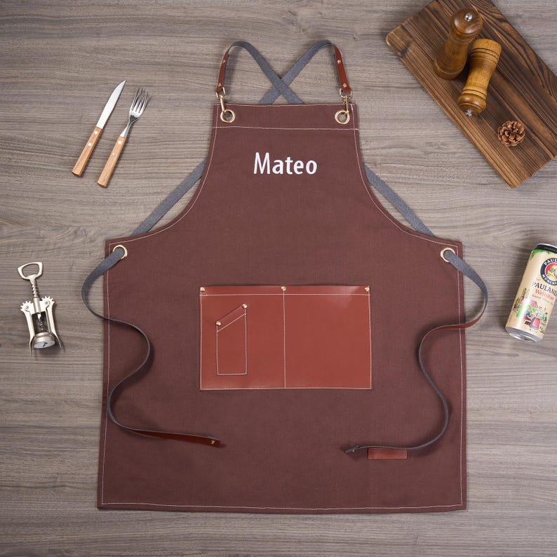 May include: A brown apron with the name "Mateo" in white, featuring a brown leather pocket and straps. The apron is on a wooden surface, with a fork, knife, and corkscrew nearby. A beer can and salt and pepper shakers are also visible.