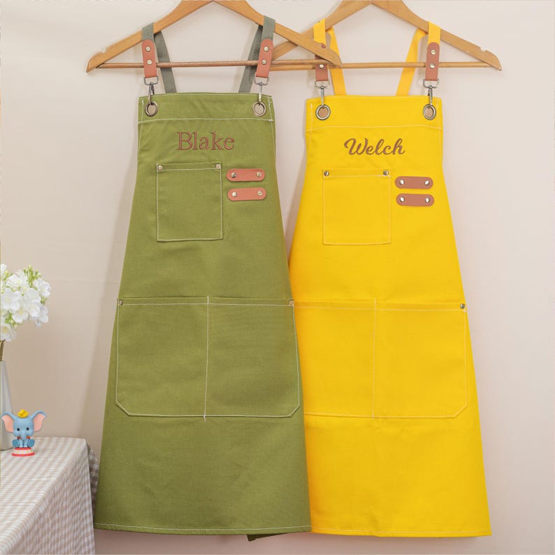 May include: Two aprons hanging on wooden hangers. One is olive green with the name "Blake" embroidered on the chest. The other is yellow with the name "Welch". Both aprons have pockets and brown leather accents.