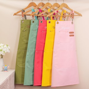 May include: Five colorful aprons hanging on wooden hangers. The aprons are in shades of green, teal, red, yellow, and pink. Each apron has a large front pocket and adjustable straps. The pink apron has the name "Lynne" embroidered on it.