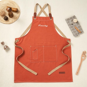 May include: An orange apron embroidered with the name "Timothy" in white. It features two front pockets, tan straps, and leather details. A small brown label is sewn on the lower right side.