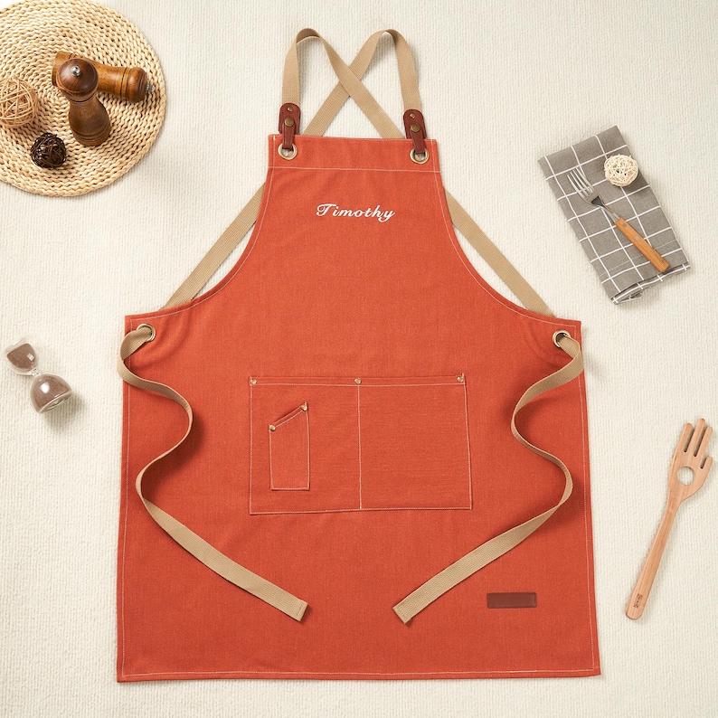 May include: An orange apron embroidered with the name "Timothy" in white. It features two front pockets, tan straps, and leather details. A small brown label is sewn on the lower right side.