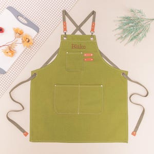 May include: Olive green apron with gray straps and accents. Features two front pockets and a chest pocket. The name "Blake" is embroidered on the chest. The straps have brown leather-like accents. A stylish and practical kitchen accessory.
