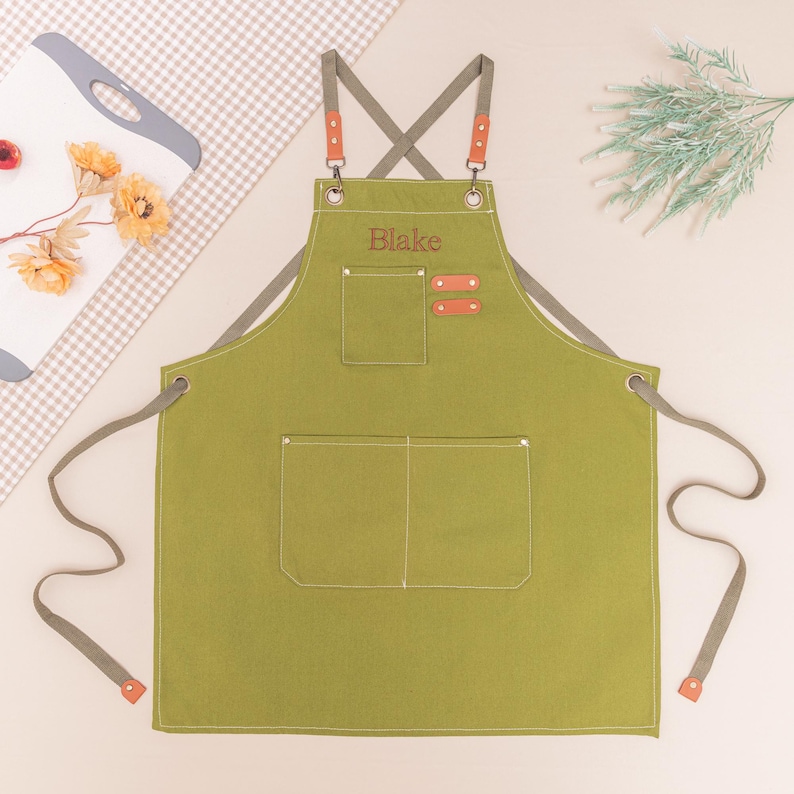 May include: Olive green apron with gray straps and accents. Features two front pockets and a chest pocket. The name "Blake" is embroidered on the chest. The straps have brown leather-like accents. A stylish and practical kitchen accessory.