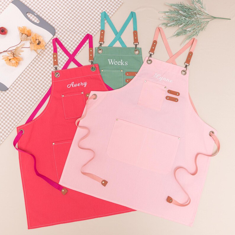 May include: Three colorful aprons: red, pink, and green. Each apron has a front pocket, adjustable straps, and a personalized name embroidered on the front. The aprons are designed for kitchen or craft use.