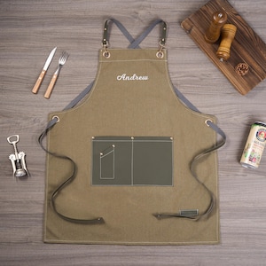 May include: An olive green apron with the name "Andrew" embroidered in white. The apron has a gray neck strap and leather straps. A knife, fork, and corkscrew are visible. A wooden cutting board, salt and pepper shakers, and a beer can are also present.