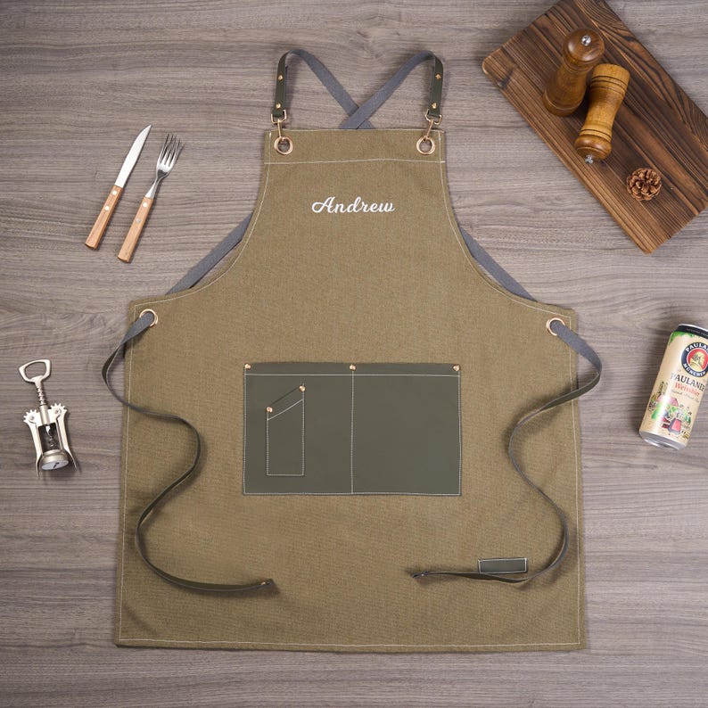 May include: An olive green apron with the name "Andrew" embroidered in white. The apron has a gray neck strap and leather straps. A knife, fork, and corkscrew are visible. A wooden cutting board, salt and pepper shakers, and a beer can are also present.