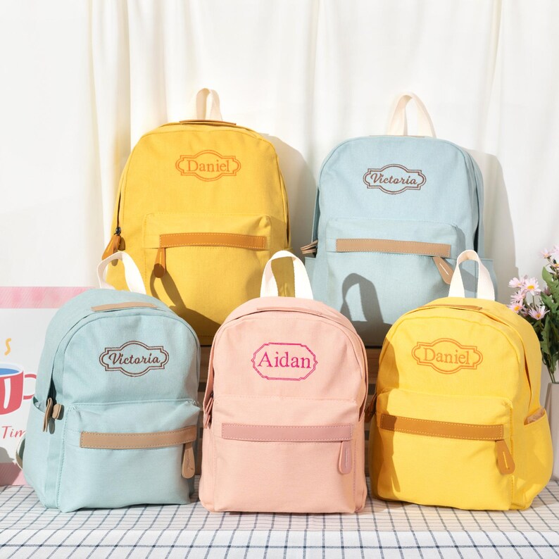 Puede incluir: Cinco mochilas de lona en amarillo, azul claro y rosa, cada una con un bolsillo frontal con cremallera, un asa superior y un nombre personalizado bordado en un marco decorativo en la parte delantera. Los nombres incluyen Daniel, Victoria y Aidan.