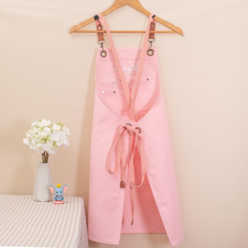 May include: A pink apron with a split front, hanging on a wooden hanger. The apron has brown leather straps and metal eyelets. A floral design is embroidered on the chest. The apron is tied at the waist with long pink ties.