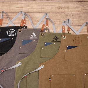 Peut inclure: Rang&eacute;e de tabliers personnalis&eacute;s noirs, gris, vert olive et marron. Chaque tablier a une poche, un nom et un motif sur le th&egrave;me du chef. Les tabliers ont des sangles grises avec des d&eacute;tails en cuir et en m&eacute;tal, parfaits pour cuisiner.