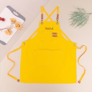 May include: A bright yellow apron with brown leather accents and the name "Welch" embroidered on the front. The apron has two large front pockets and adjustable straps. The apron is made of a durable fabric.