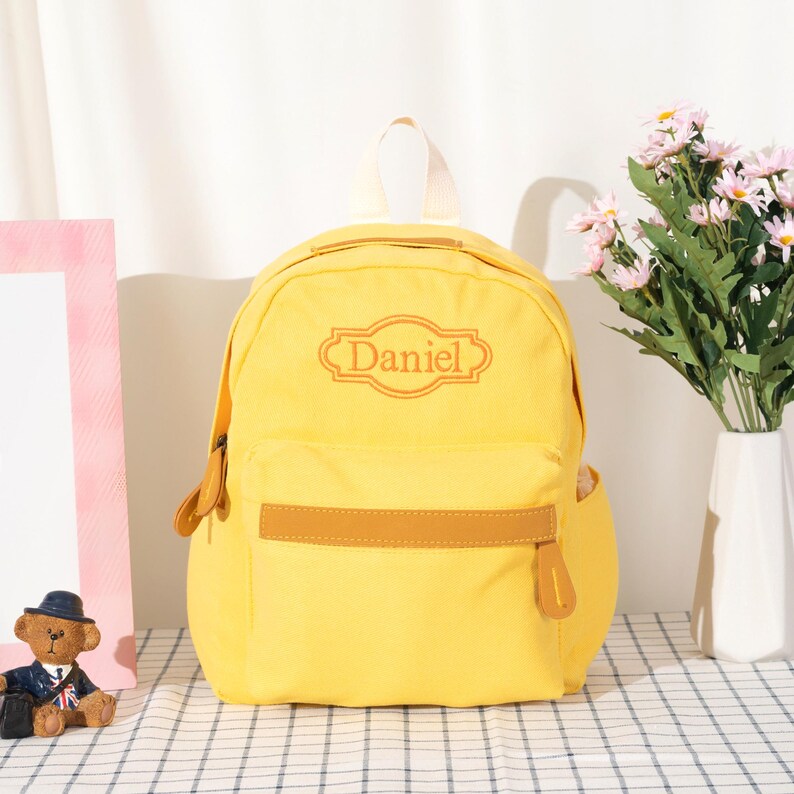 Puede incluir: Una mochila de color amarillo brillante con un asa superior y un bolsillo frontal con cremallera que incluye una franja horizontal marr&oacute;n. El nombre Daniel est&aacute; bordado en un marco decorativo naranja en la parte superior frontal de la mochila.