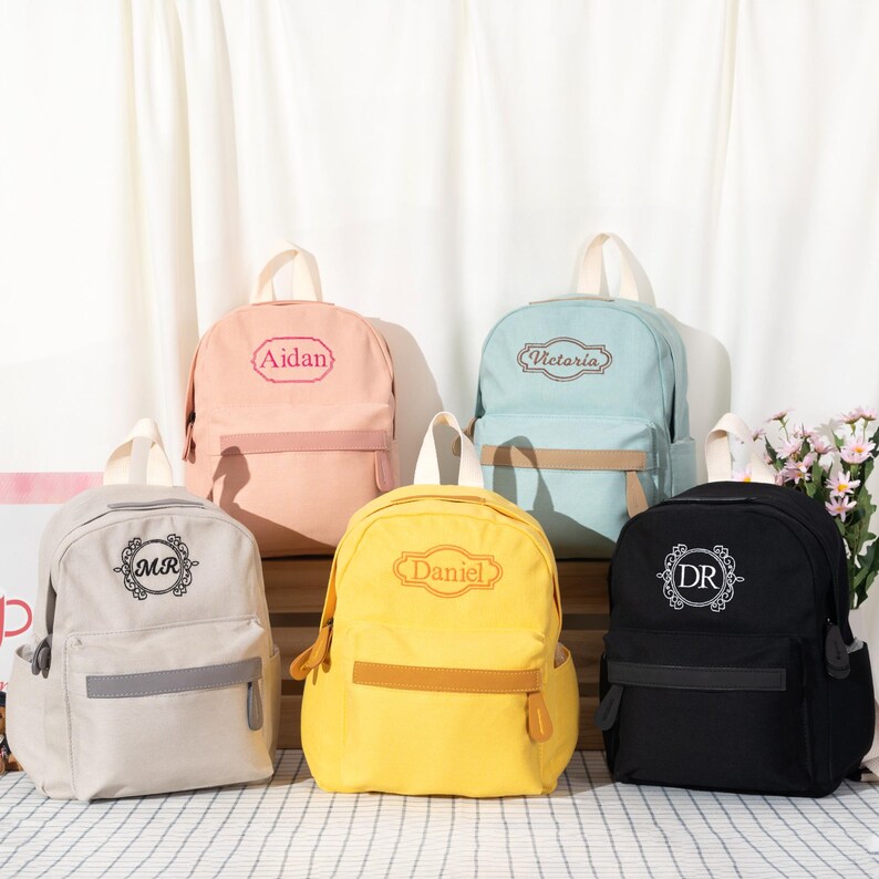 Puede incluir: Cinco mochilas en beige, rosa, azul claro, amarillo y negro, cada una con bolsillo frontal, asa superior y bordado personalizado. La bolsa beige muestra MR, la rosa dice Aidan, la azul claro Victoria, la amarilla Daniel y la negra DR.