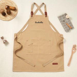 May include: A beige apron embroidered with the name "Isabella" in black. The apron features a pocket, leather details, and adjustable straps. Kitchen utensils and accessories, including a wooden fork and spoon, are arranged around it.