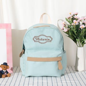 Puede incluir: Una mochila de lona azul claro con un asa superior y un bolsillo frontal con cremallera y ribete de piel sint&eacute;tica beige. El nombre Victoria est&aacute; bordado en letra cursiva marr&oacute;n dentro de un borde decorativo en la parte superior frontal.