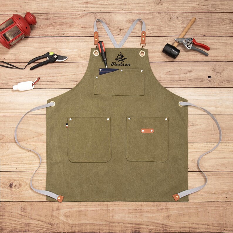 Peut inclure: Un tablier vert olive avec des sangles grises et des d&eacute;tails en cuir. Le tablier comporte plusieurs poches et le nom "Hudson" est imprim&eacute; au-dessus d'un motif de flamme. Des outils et une lanterne rouge sont dispos&eacute;s autour du tablier sur une surface en bois.